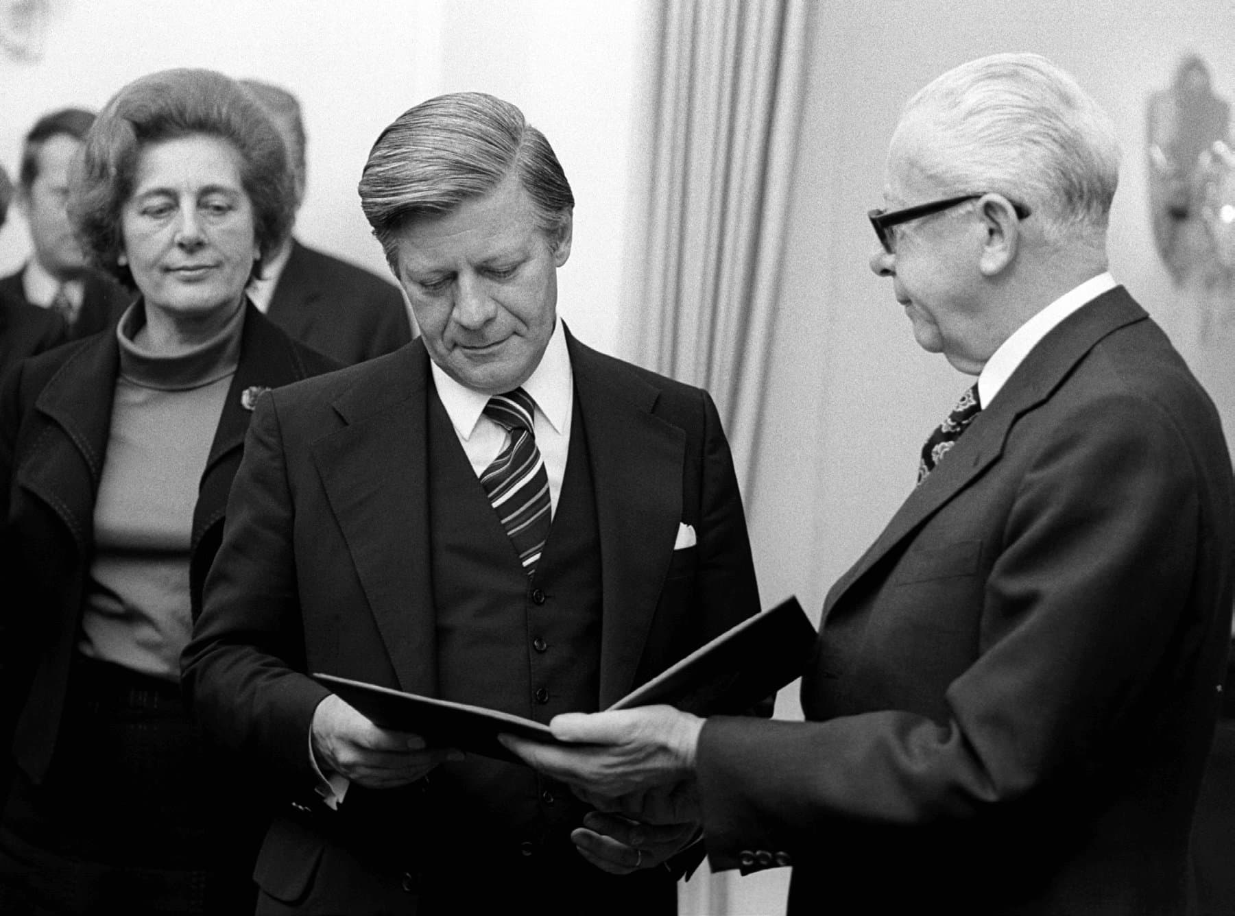 A black-and-white photo depicts Helmut Schmidt looking at the certificate of appointment in his hands. Federal President Gustav Heinemann is standing to his right at looks at him while also holding that certificate.
