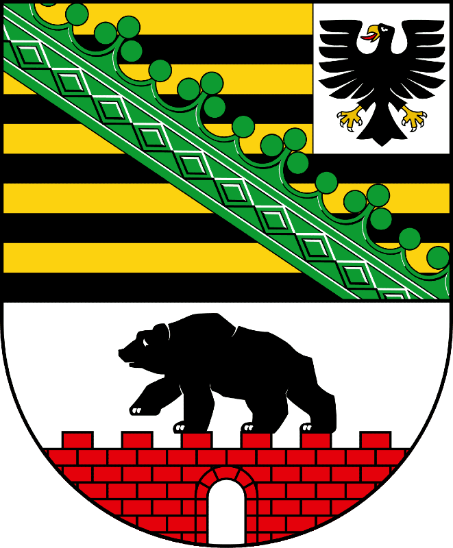 A shield with a flag on its upper part and a black bear climbing on top of the wall made of red bricks. A flag has ten horizontal stripes (yellow-black), a green ornament placed on its diagonal and a 2D black eagle on the white background in the top-right corner.